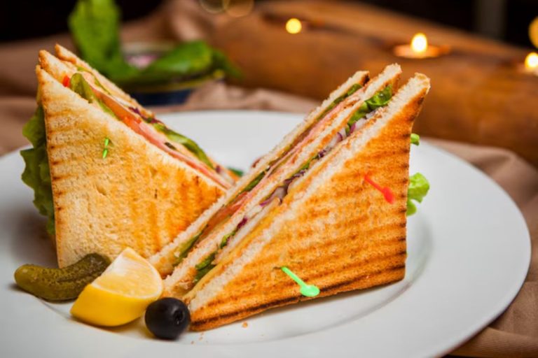 A Toastul close-up of a grilled club sandwich filled with fresh vegetables, cheese, and meat, served with pickles, lemon, and an olive on a white plate.