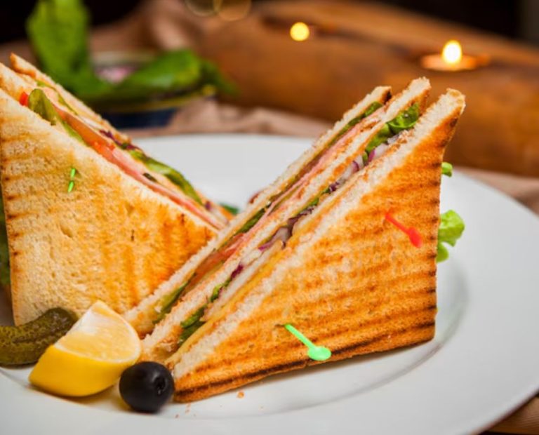 A Toastul close-up of a grilled club sandwich filled with fresh vegetables, cheese, and meat, served with pickles, lemon, and an olive on a white plate.